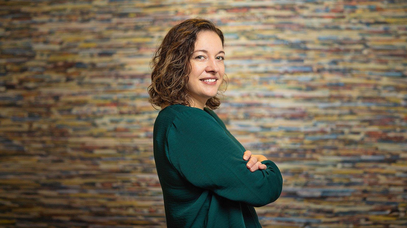 Aron Gelineau stands with her arms crossed and smiles at the camera. 她有一头棕色的卷发，穿着一件蓝绿色的毛衣.