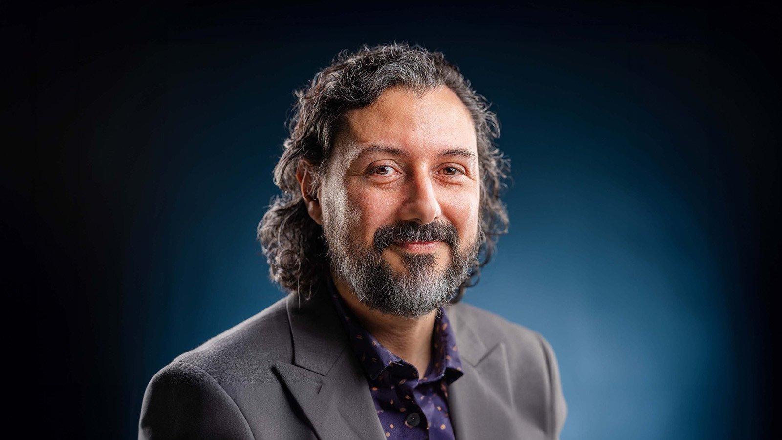 Plinio Pinto smiles at the camera. He has curly brown hair and a beard streaked with gray, and he wears a gray blazer.