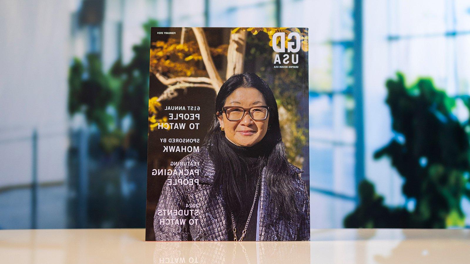 A copy of “Graphic Design USA” magazine depicting a woman wearing glasses and a jacket stands atop a white table.