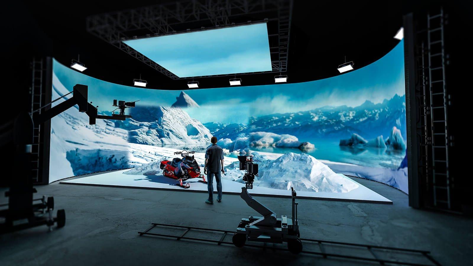 A man wearing jeans, sneakers, a plaid shirt stands in a production studio. He is facing a large LED screen showing an icy mountain range and a snowmobile.