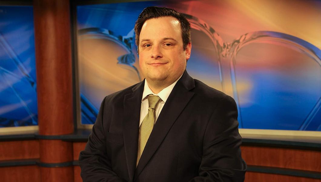 Dan Hanson sits at a news desk in a television studio. He is smiling and wearing a black suit with a yellow tie.