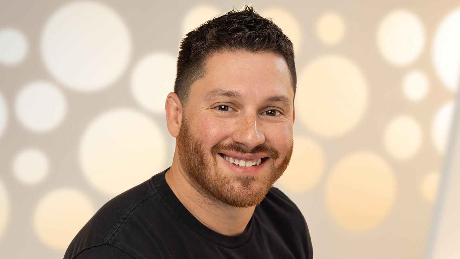 A headshot of a man with short brown hair and a black t-shirt smiling against a white background.