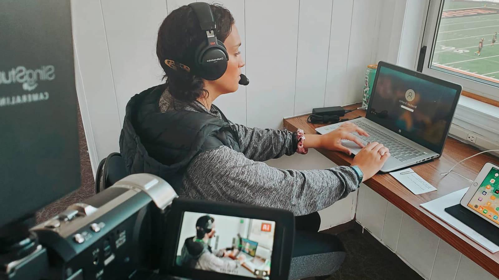 毕业生悉尼菲利普斯, a woman with dark brown hair wearing a grey hooded sweatshirt and black over-ear headphones, is seated at a desk on her laptop in a press box overlooking athletes in uniform on a football field.