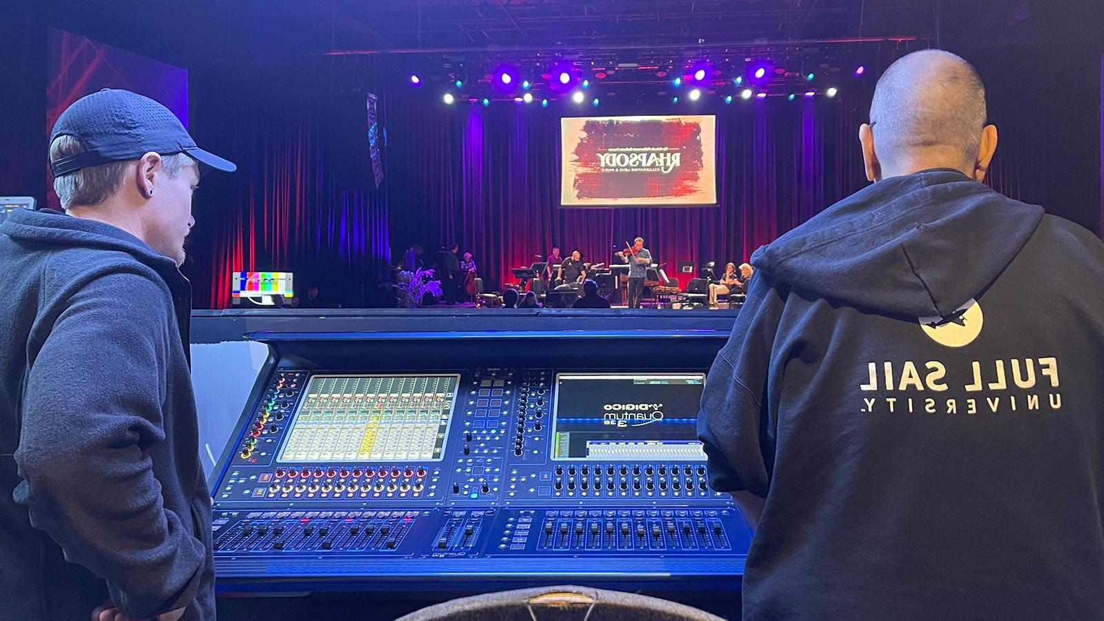 Two people's backs are facing the camera as they overlook an audio console and stage at The Plaza Live.