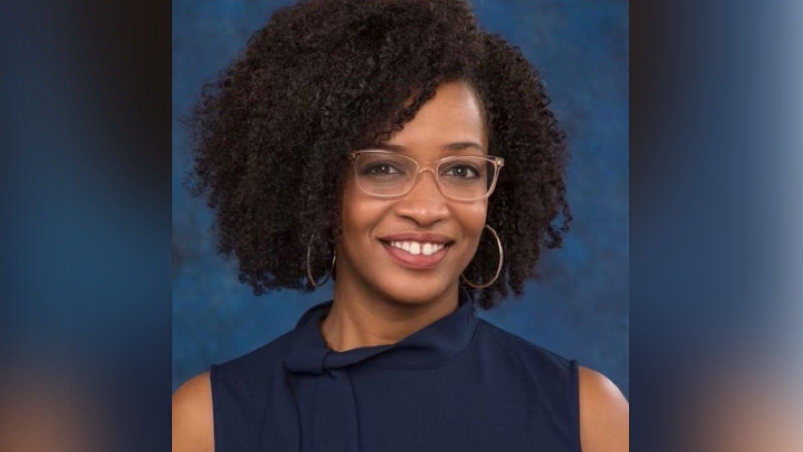 Tiana, an African American woman, sits smiling at the camera. 她穿着一件无袖的藏青色上衣, 银环耳环, 还有半透明米色镜框的眼镜.