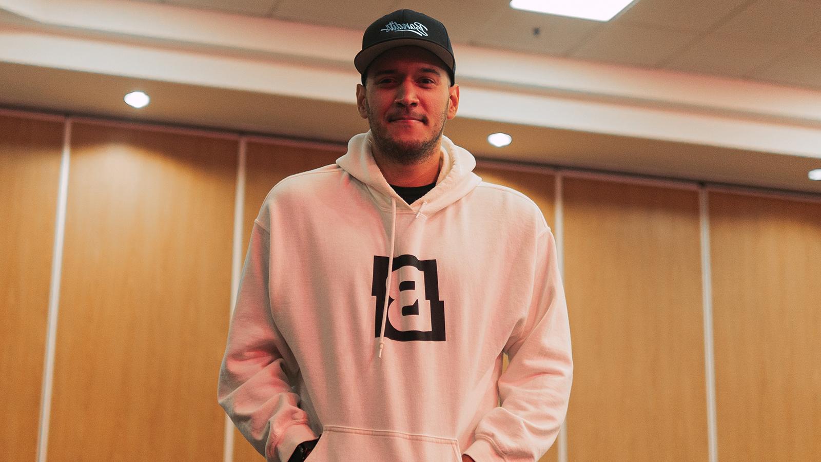 A man standing against a wood paneled wall in a white hooded sweatshirt featuring the Bandits logo and a black 强盗的游戏 snapback cap.