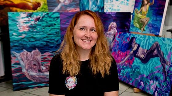 Jessica sits in front of several of her colorful paintings. She is wearing a black t-shirt and is smiling.