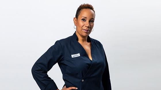 Grad Elle Simone Scott standing and smiling against a white backdrop while wearing a blue double-breasted chef's jacket with an America's Test Kitchen patch on the breast.