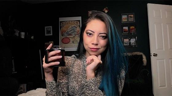 A woman with long brown hair with blue highlights holding a mug while seated in a home office.