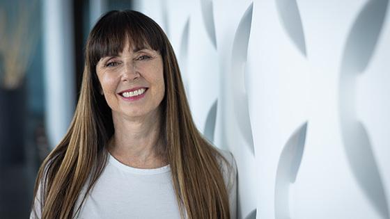 A woman with long brown hair and bangs smiling while standing against a geometric white backdrop.