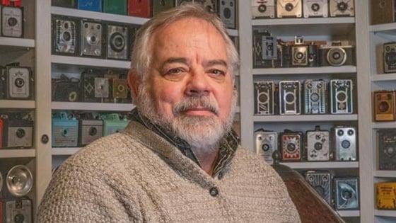 Randy sits in front of a wall covered by shelves of vintage cameras. He is wearing a light brown sweater.