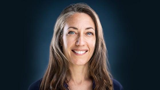 Kelly sits in front of a blue background wearing a blue blouse. She has blue eyes and is smiling.