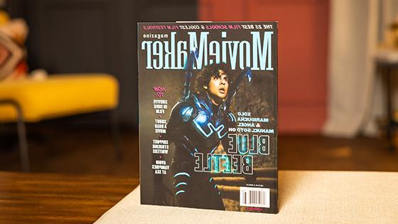 A copy of MovieMaker Magazine sits on a table. An actor in a Blue Beetle costume is on the cover. The headline “The 25 Best Film Schools & Coolest Film Festivals” runs across the top.