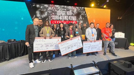 Techfest tournament winners holding large scholarship checks on stage alongside Full Sail staff members in the Fortress.