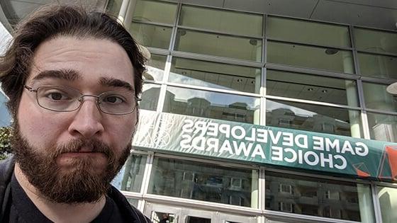 Ryan Miller pictured from above the shoulders in a black leather jacket with silver wireframe glasses in front of a sign reading “Game Developers Choice Awards.”