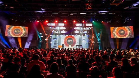 A person wearing a suit speaks on a stage in front of a large crowd. There is a large screen with a logo that says 名人堂 XII behind them. 红色、蓝色和白色的灯照亮了房间.