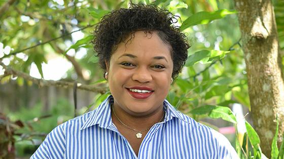 Jessica stands in front of a lush garden, surrounded by greenery. She is wearing a blue pinstripe shirt and is smiling.