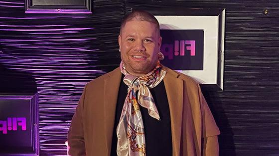 Javi Simons stands in front of a purple wall. He wears a black t-shirt, jeans, a brown overcoat, and a colorful scarf. He is smiling.