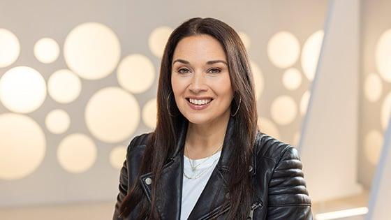 A headshot of a woman with straight brown hair and hoop earrings smiling while wearing a leather motorcycle jacket against a white background.