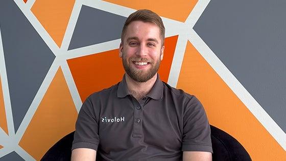 A man with light hair and a beard wearing a 灰色 Holovis polo while seated and smiling in front of an orange, 灰色, 白色几何背景.