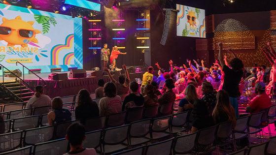 Several students raise their hands attentively at the Summer Sizzle as two staff members call on participants. The large room is colorfully lit up and looks very lively.