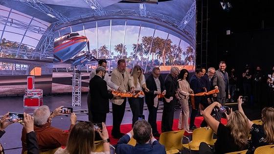 A group of eight men and two women stand on a red carpet in front of a large screen displaying an airplane and palm trees. They are each holding a pair of scissors and cutting a long orange ribbon. The seven audience members in front of them are photographing them with cell phones.