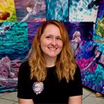 Jessica sits in front of several of her colorful paintings. She is wearing a black t-shirt and is smiling.