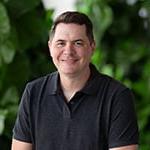 A man with short brown hair is wearing a dark grey polo while smiling against a lush green backdrop.