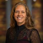 Phoebe Elefante, a woman with shoulder-length light brown hair and a black blouse smiling in front of an out-of-focus brown backdrop.