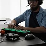 A man wearing a gaming headset sits at a desk with a gaming keyboard, an Xbox controller, and a Dungeons & Dragons die.