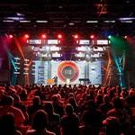 A person wearing a suit speaks on a stage in front of a large crowd. 的re is a large screen with a logo that says 名人堂 XII behind them. Red, blue, and white lights illuminate the room.