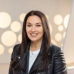 A headshot of a woman with straight brown hair and hoop earrings smiling while wearing a leather motorcycle jacket against a white background.