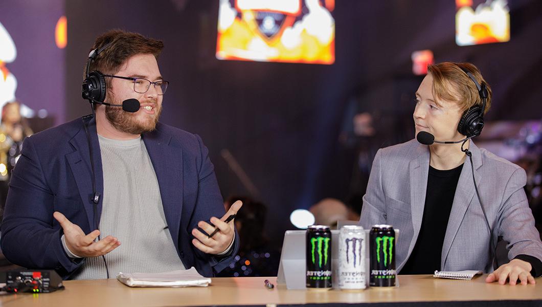 Two well-dressed people are seated at a hosting desk while wearing headsets and chatting.