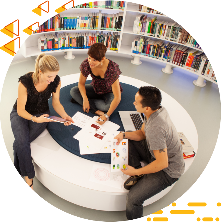 Aerial photo of three 学生 collaborating on design work while seated on a large ottoman, 书籍是背景.